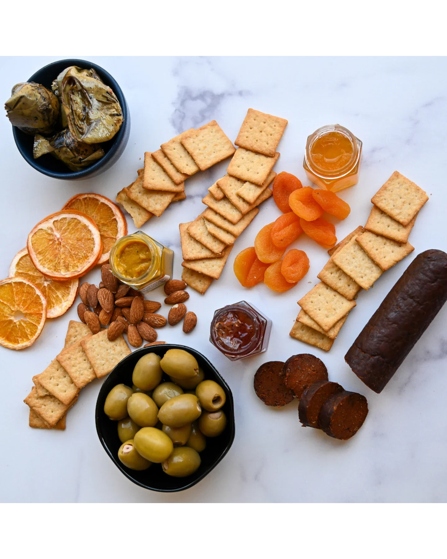 assorted dried fruit, crackers, olives, vegan salami, and jam on a white marble background
