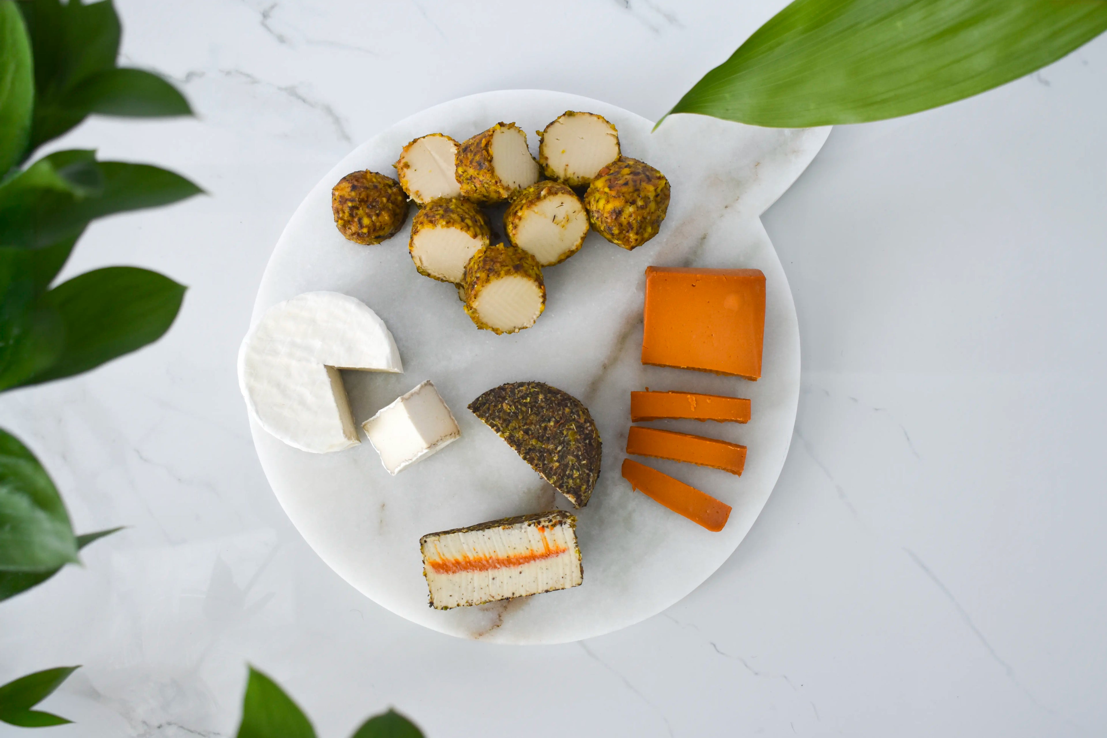 An overhead flat-lay of assorted cheeses arranged on a round white marble serving board with a handle, set on a light marble surface with green leaves at the edges. The cheeses include soft white wheels and cubes, orange cheddar-like slices, a dark rind cheese half, and a sliced cheese showing cream and orange layers.
