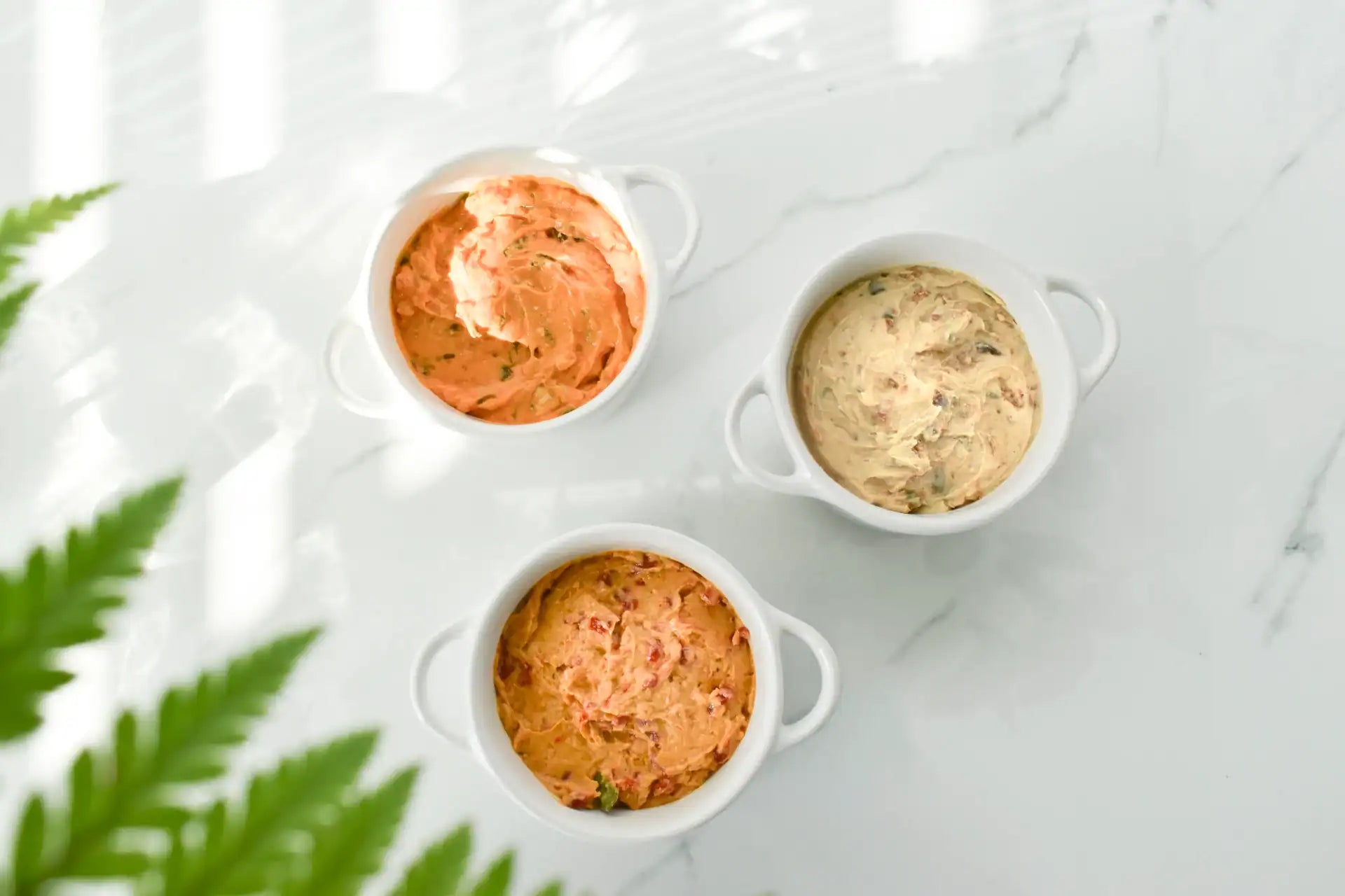 Three small white ceramic ramekins filled with creamy dairy-free dips and spreads, arranged on a light marble surface with soft shadows and green leaves in the corner.