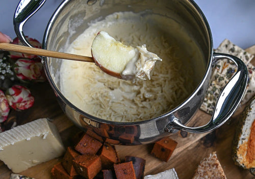 Person dipping an apple into a bowl of cheese fondue with a charcuterie board in the background.