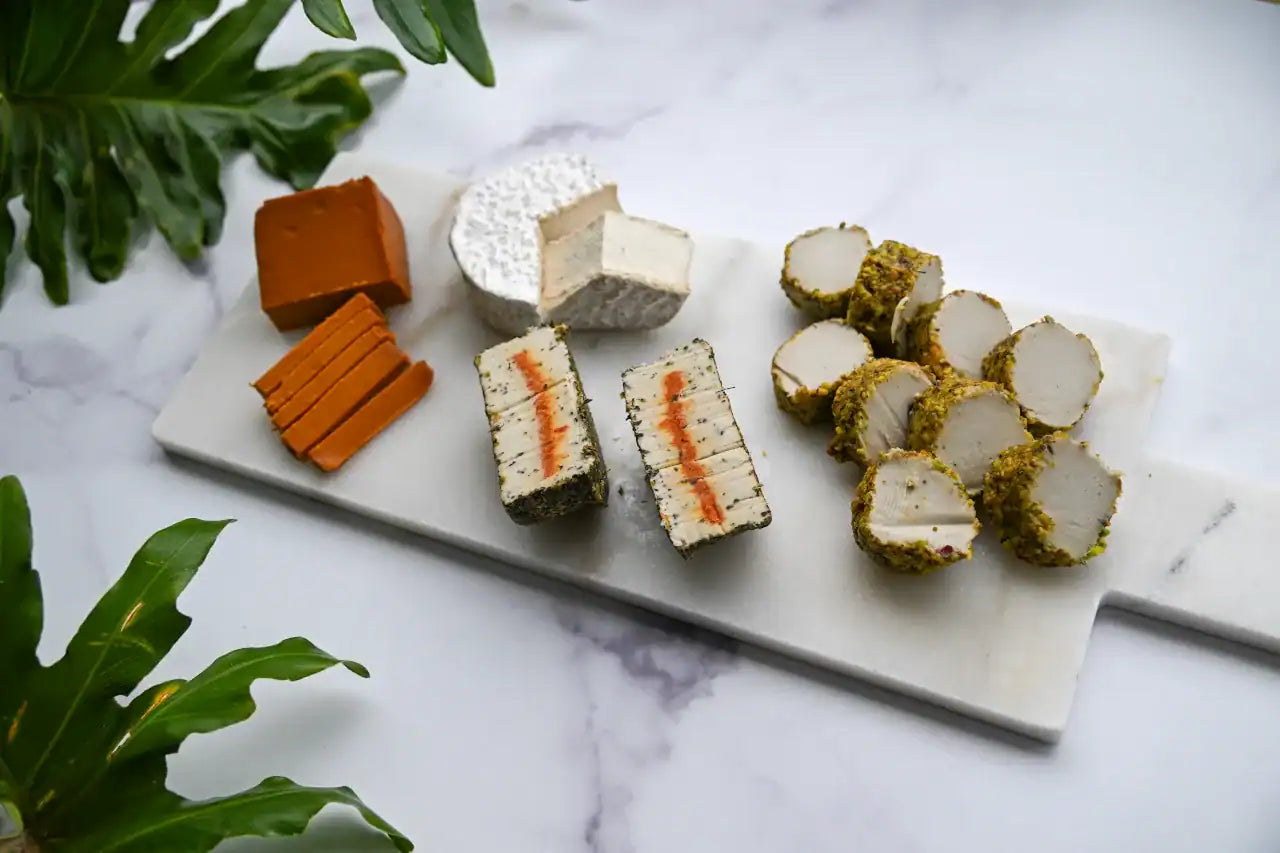 Assorted vegan cheese products on a marble board with green leaves in the background.
