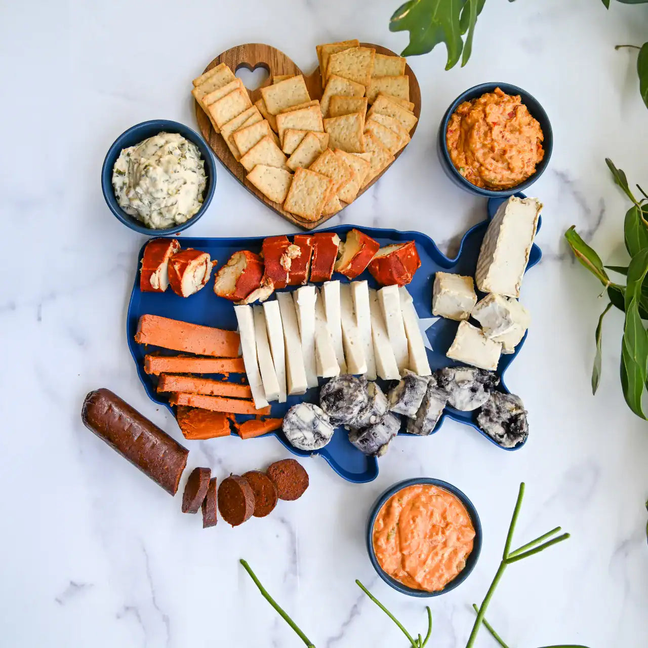 Assorted meats, cheeses, and crackers on a heart-shaped platter with dips on a marble surface.