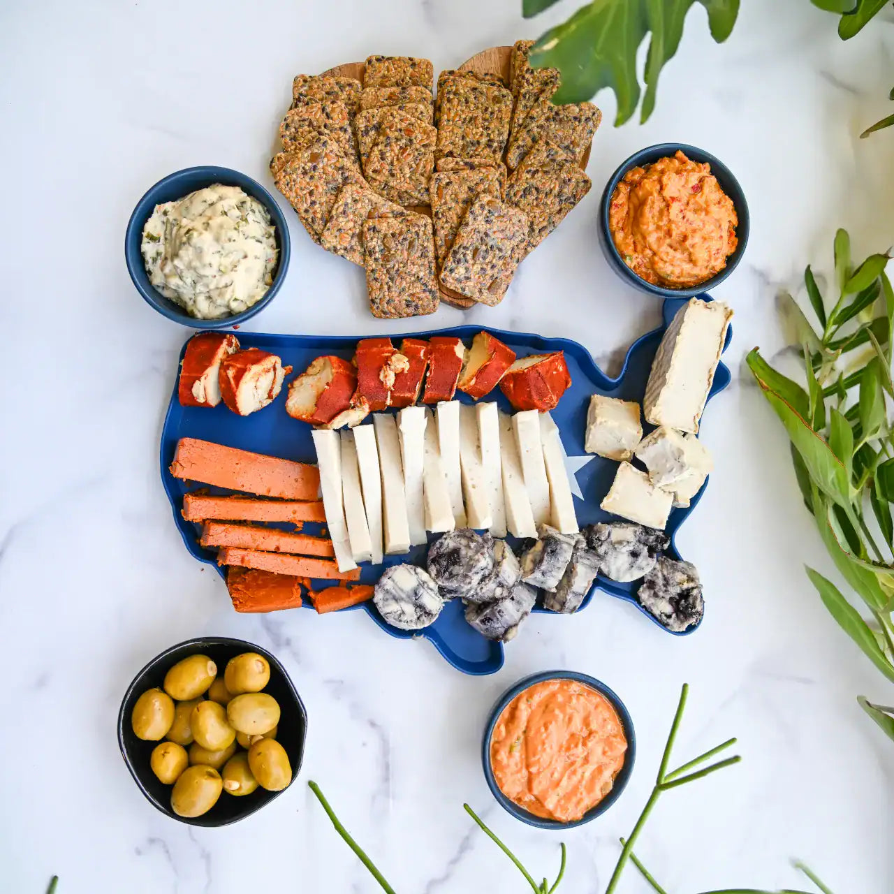 Platter of assorted cheeses, crackers, and dips on a white marble surface with greenery.