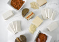 An overhead flat-lay display of various dairy-free cheeses and vegan cured meats on a white marble surface. The assortment includes white and yellow cheeses, sliced cheeses with orange-red fillings, dark-rind cheeses, shredded cheese, and bowls containing reddish-brown crumbled topping and sliced pepperoni.