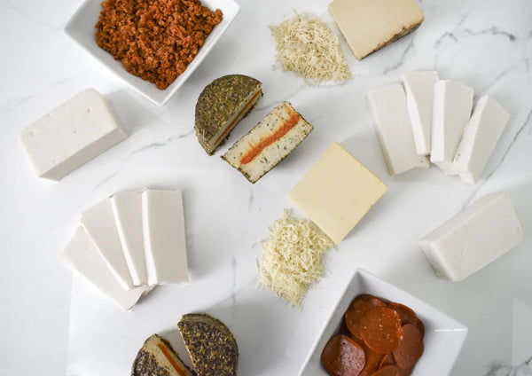 An overhead flat-lay display of various dairy-free cheeses and vegan cured meats on a white marble surface. The assortment includes white and yellow cheeses, sliced cheeses with orange-red fillings, dark-rind cheeses, shredded cheese, and bowls containing reddish-brown crumbled topping and sliced pepperoni.
