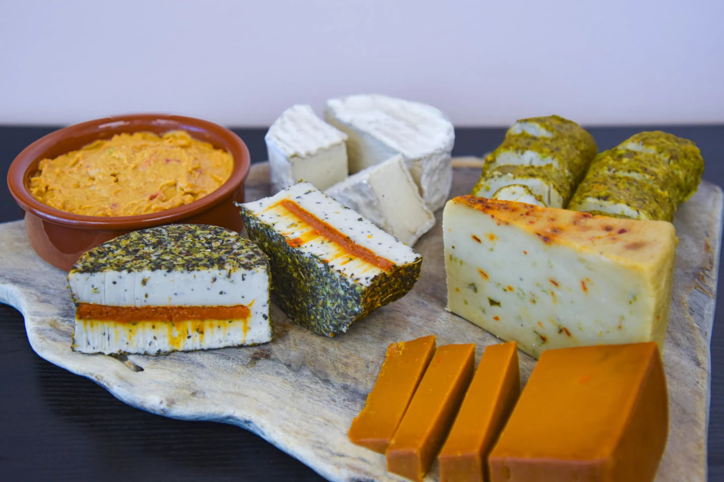 Assorted types of cheese on a wooden board with a bowl of pimento cheese.