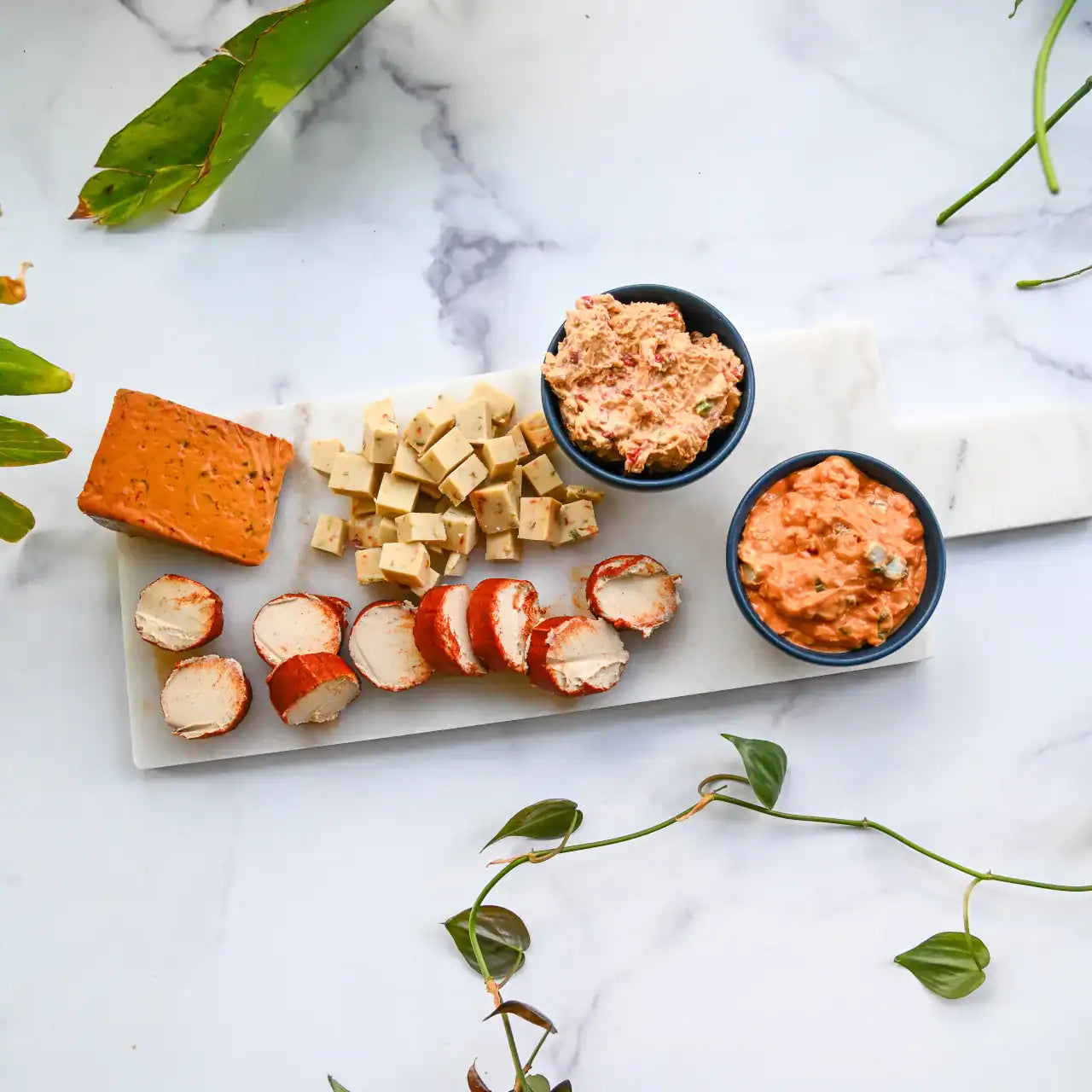 Platter with assorted dairy-free cheese and two small bowls of dip on a marble surface.