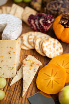 An assortment of Pepper Jack and Brie cheeses, crackers, persimmon slices, grapes, and nuts arranged on a wooden board in a close-up food scene.