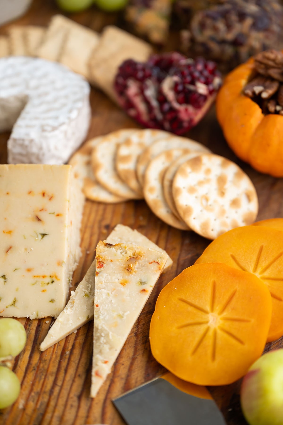 An assortment of Pepper Jack and Brie cheeses, crackers, persimmon slices, grapes, and nuts arranged on a wooden board in a close-up food scene.