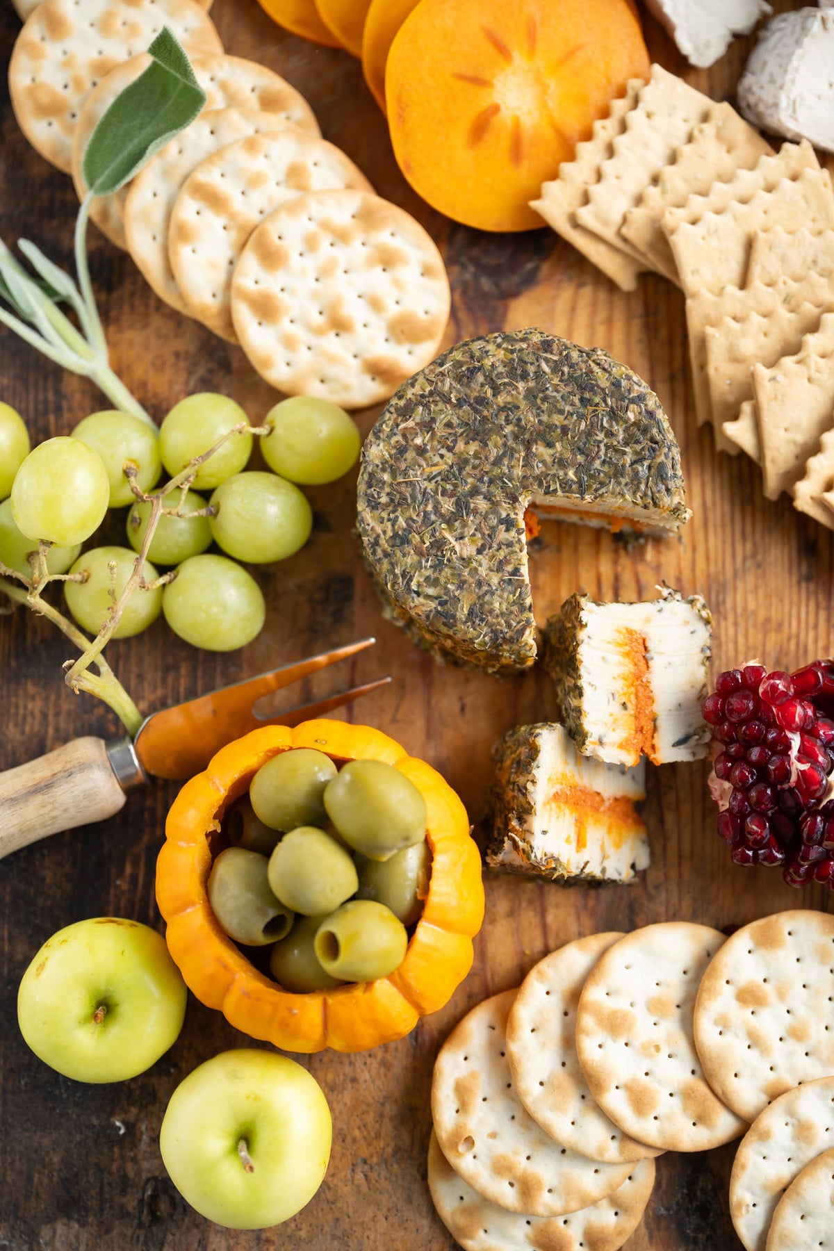 A styled charcuterie or cheese board on a wooden surface with assorted crackers, a round cheese wheel and cut pieces showing orange filling, green grapes, green olives in a small orange bowl, apples, pomegranate, and herbs.
