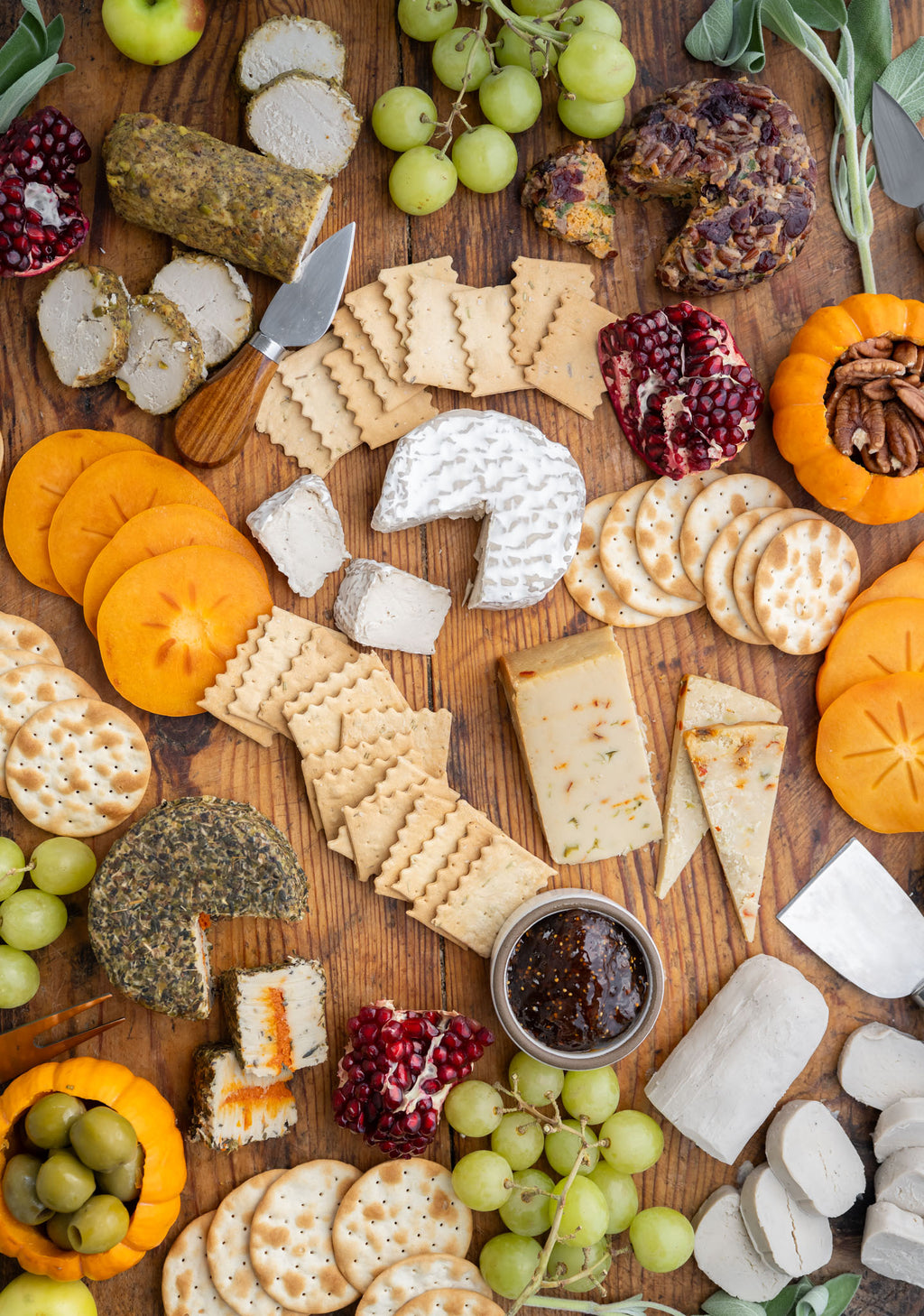 An overhead flat-lay of a gourmet cheese and charcuterie board arranged on a wooden surface, featuring various cheeses, crackers, grapes, persimmon, pomegranate, nuts, olives, and a small jar of dark spread.