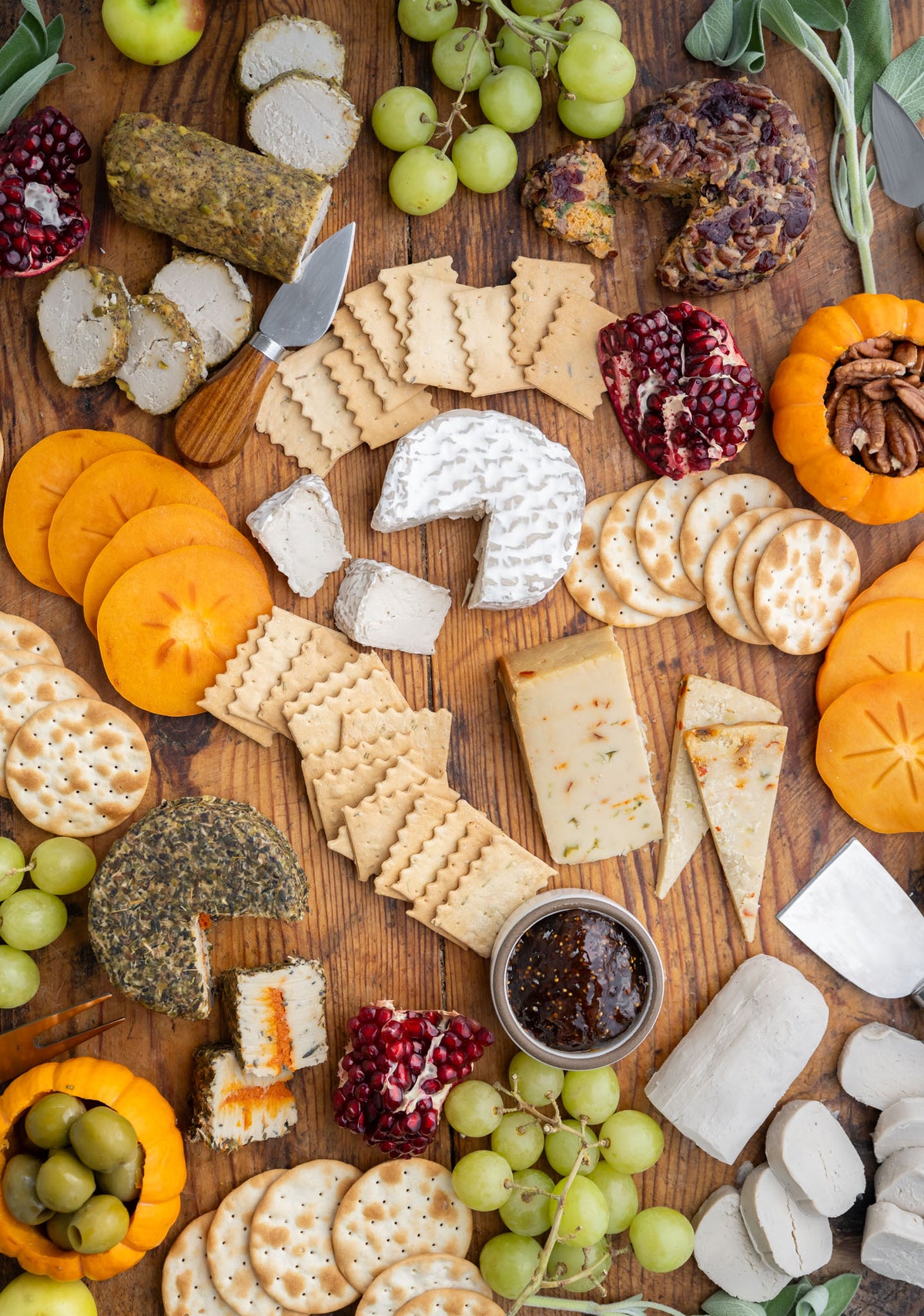 An overhead flat-lay of a gourmet cheese and charcuterie board arranged on a wooden surface, featuring various cheeses, crackers, grapes, persimmon, pomegranate, nuts, olives, and a small jar of dark spread.