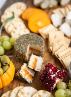A close-up, top-down view of a rustic charcuterie board featuring A Tomato Herb Fromage cheese wheel with a slice cut out, surrounded by crackers, grapes, olives, pomegranate, and orange slices on a wooden board.