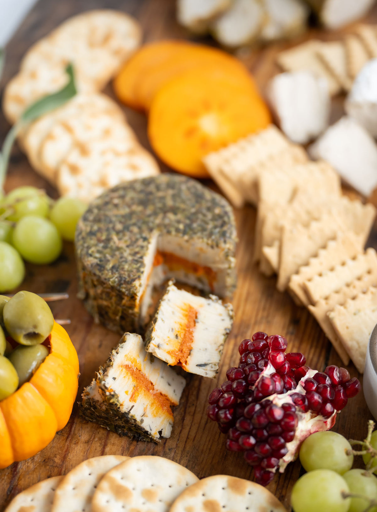 A close-up, top-down view of a rustic charcuterie board featuring A Tomato Herb Fromage cheese wheel with a slice cut out, surrounded by crackers, grapes, olives, pomegranate, and orange slices on a wooden board.