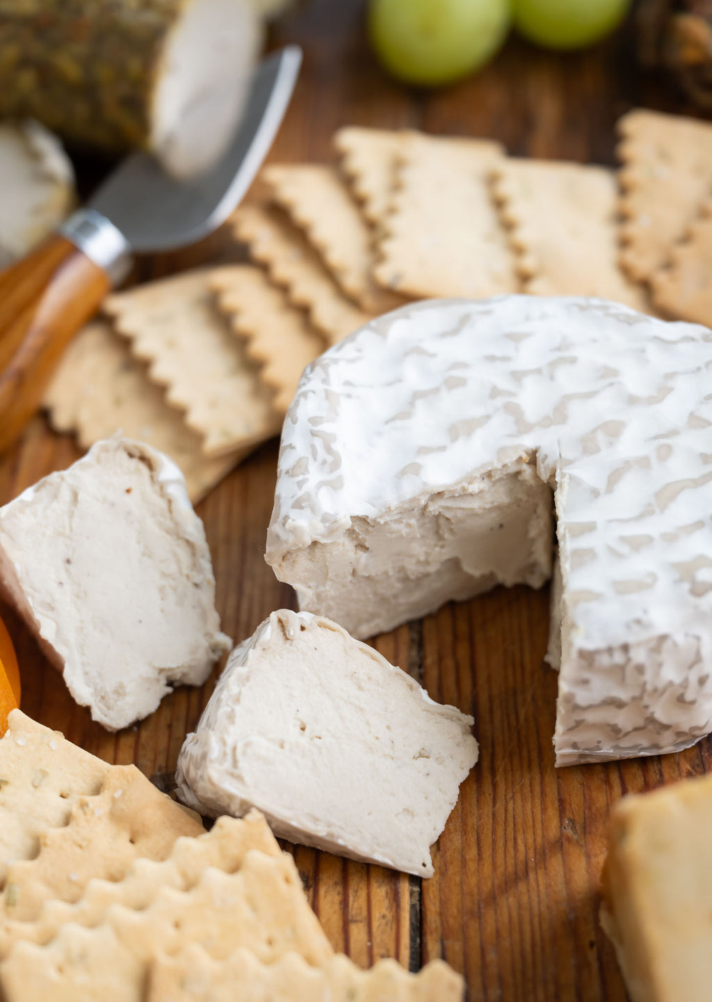 A wheel of soft Brie cheese with a textured rind, shown sliced on a wooden board with crackers, grapes, and a cheese knife.