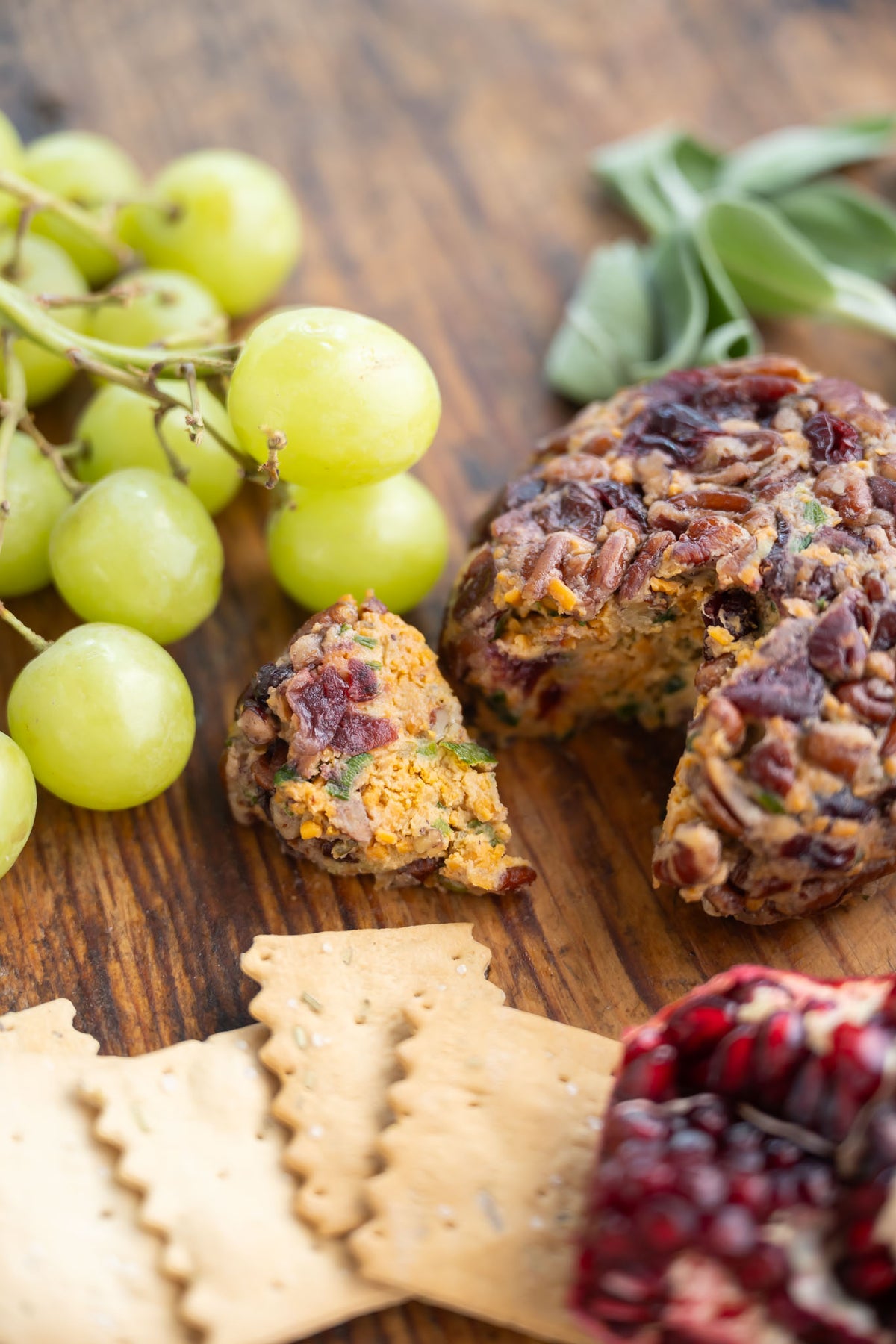A close-up food scene showing slice of Rebel Cheese seasonal Cranberry Pecan Cheeseball on a wooden board, surrounded by green grapes, crackers, herbs, and a pomegranate.