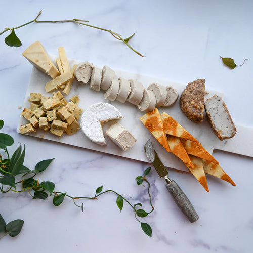 Assorted dairy-free cheeses on a marble board with a knife, surrounded by greenery. 