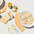 Vegan artichoke dip kit with plant-based cream cheese, dairy-free Parmesan, marinated artichokes, red wine mustard, and sourdough crackers on a white marble background