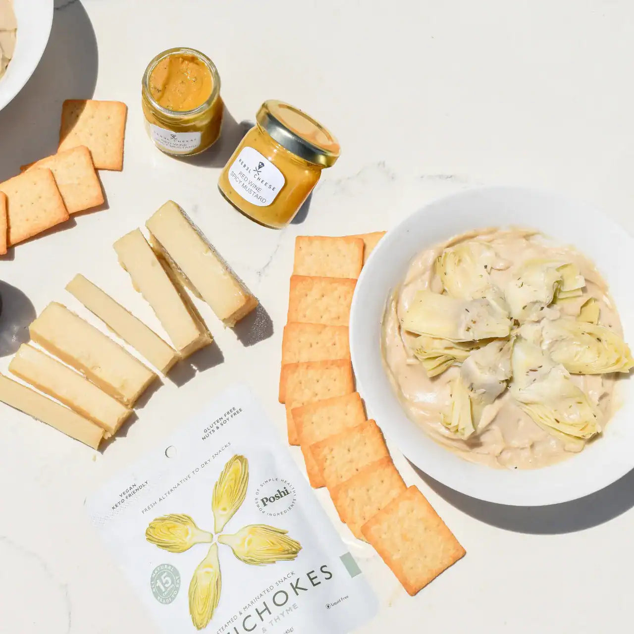 Vegan artichoke dip kit with plant-based cream cheese, dairy-free Parmesan, marinated artichokes, red wine mustard, and sourdough crackers on a white marble background