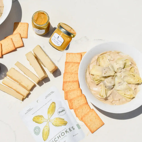 Vegan artichoke dip kit with plant-based cream cheese, dairy-free Parmesan, marinated artichokes, red wine mustard, and sourdough crackers on a white marble background
