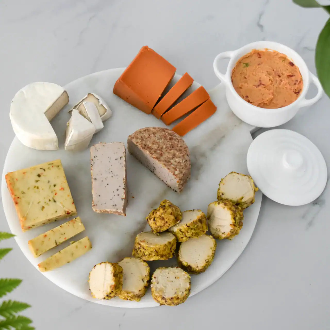 An overhead flat-lay of a charcuterie and cheese board on a white marble platter, featuring various cheeses and a small white bowl of Pimento cheese dip with its lid beside it, set on a light marble surface with green leaves at the edges.