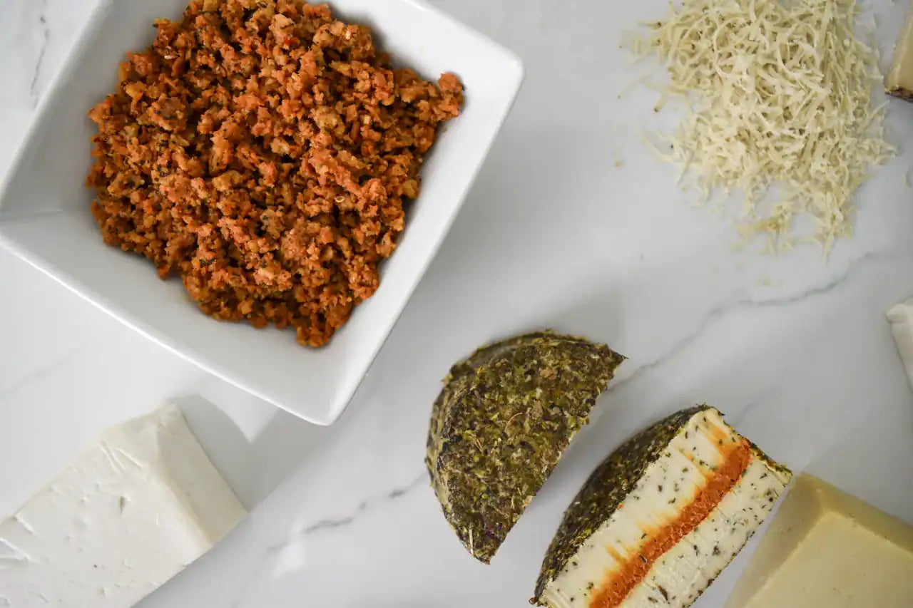 Overhead flat-lay of a food product on a white marble surface, featuring a white square bowl filled with browned ground meat, two halves of a baked cheese or cheese-based bread with herb crust and orange filling, blocks of white and yellow cheese, and a pile of shredded cheese.