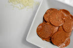 Sliced pepperoni served on a white square plate, with shredded cheese scattered in the upper left on a white surface.