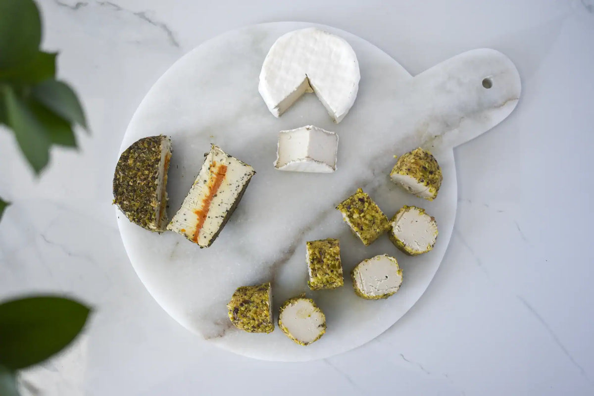 An overhead flat-lay of assorted cheeses on a round white marble serving board with a handle, placed on a light marble surface. The cheeses include a soft white cheese with a wedge removed, a sliced cheese with a cream-colored center and orange-red filling, and several round cheeses topped with green roasted pistachio coating.