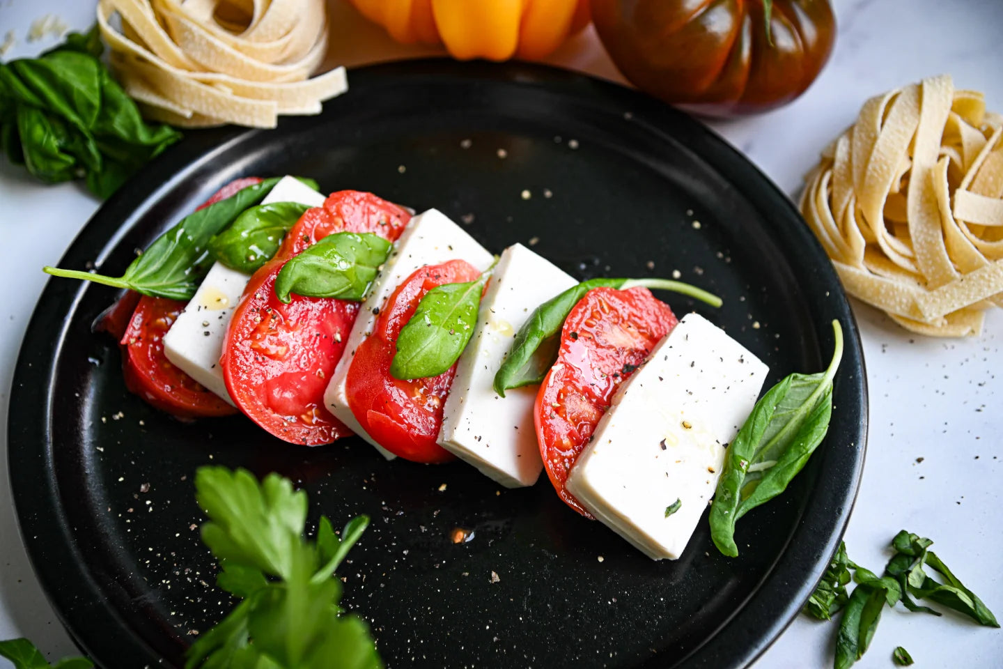 Caprese salad with mozzarella, tomatoes, and basil on a black plate with pasta and heirloom tomatoes in the background.