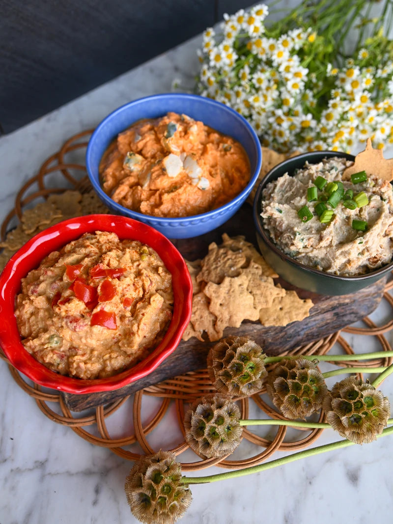 Three bowls of different dips (Pimento Cheese, Bacon Scallion Dip, and Buffalo Blue Cheese Dip) on a woven mat with flowers in the background