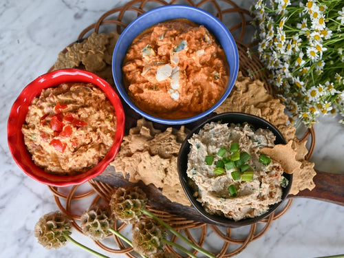 Three bowls of different dips (Pimento Cheese, Bacon Scallion Dip, and Buffalo Blue Cheese Dip) on a woven mat with flowers in the background