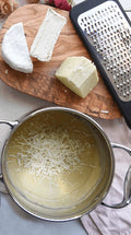 Grated cheese in a pot with a wooden board of cheese and a grater in the background.