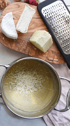 Grated cheese in a pot with a wooden board of cheese and a grater in the background.