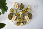 A top-down view of sliced and cubed Honee Pistachio Chèvre and Tomato Herb Fromage, arranged on a white marble serving board with a white handle, set on a white marble surface with green leaves in the corner.