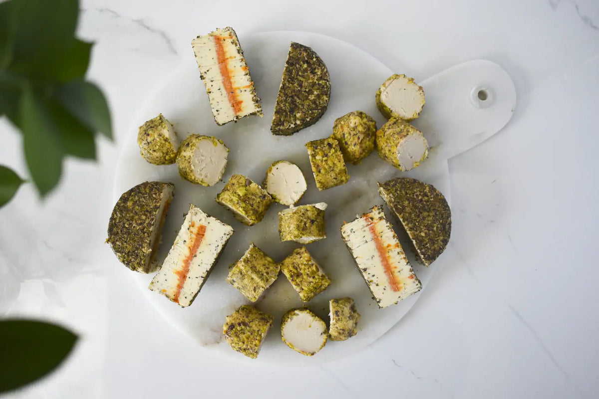 A top-down view of sliced and cubed Honee Pistachio Chèvre and Tomato Herb Fromage, arranged on a white marble serving board with a white handle, set on a white marble surface with green leaves in the corner.