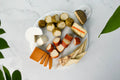 An overhead flat-lay of a white marble serving board filled with assorted dairy-free cheeses on a light gray marble surface. Visible items include several cheeses in different textures and colors, including a white semi-hard cheese, orange cheddar-like slices, pale yellow cheese wedges, and greenish-brown rind cheeses.