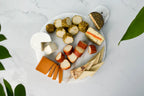 An overhead flat-lay of a white marble serving board filled with assorted dairy-free cheeses on a light gray marble surface. Visible items include several cheeses in different textures and colors, including a white semi-hard cheese, orange cheddar-like slices, pale yellow cheese wedges, and greenish-brown rind cheeses.