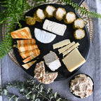 Assorted dairy-free cheeses on a black plate with crackers and a small bowl of spreadable food, surrounded by greenery.