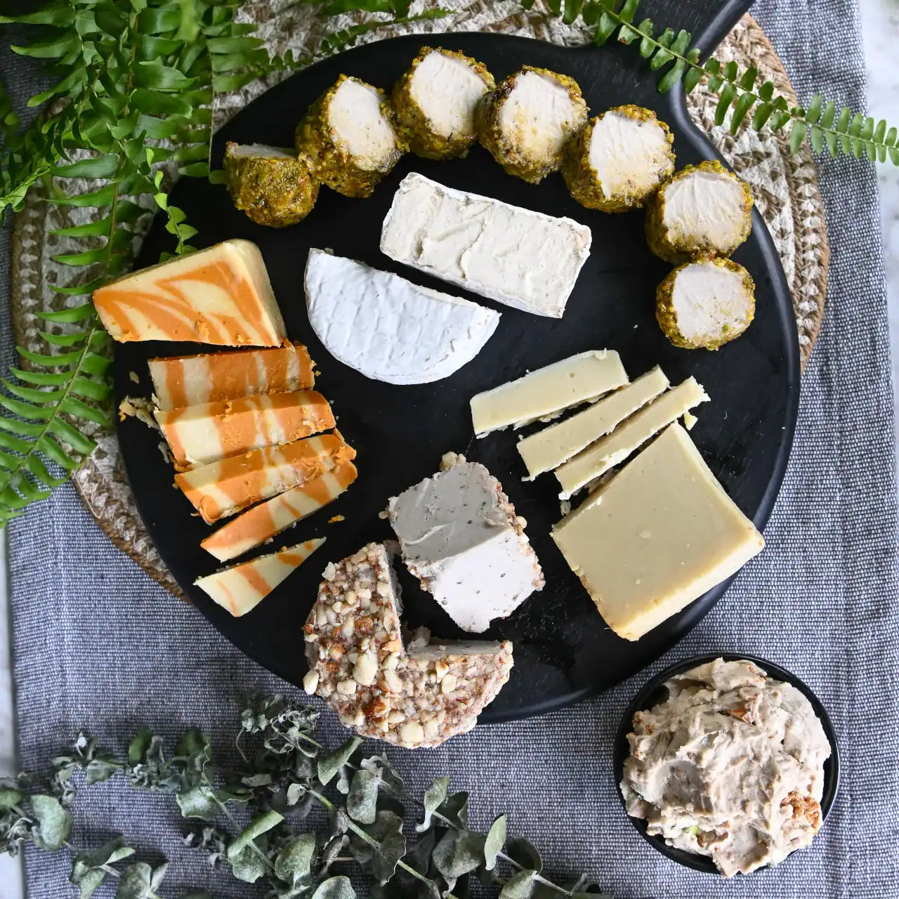 Assorted dairy-free cheeses on a black plate with crackers and a small bowl of spreadable food, surrounded by greenery.