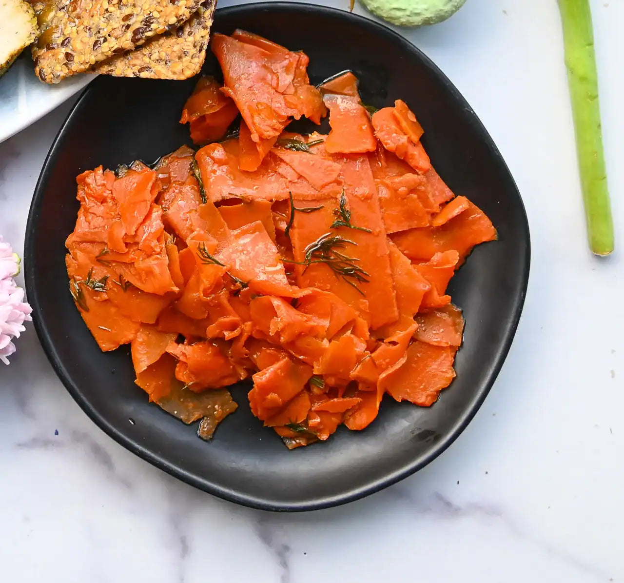Smoked carrot lox on a black plate with crackers and vegetables on a marble surface