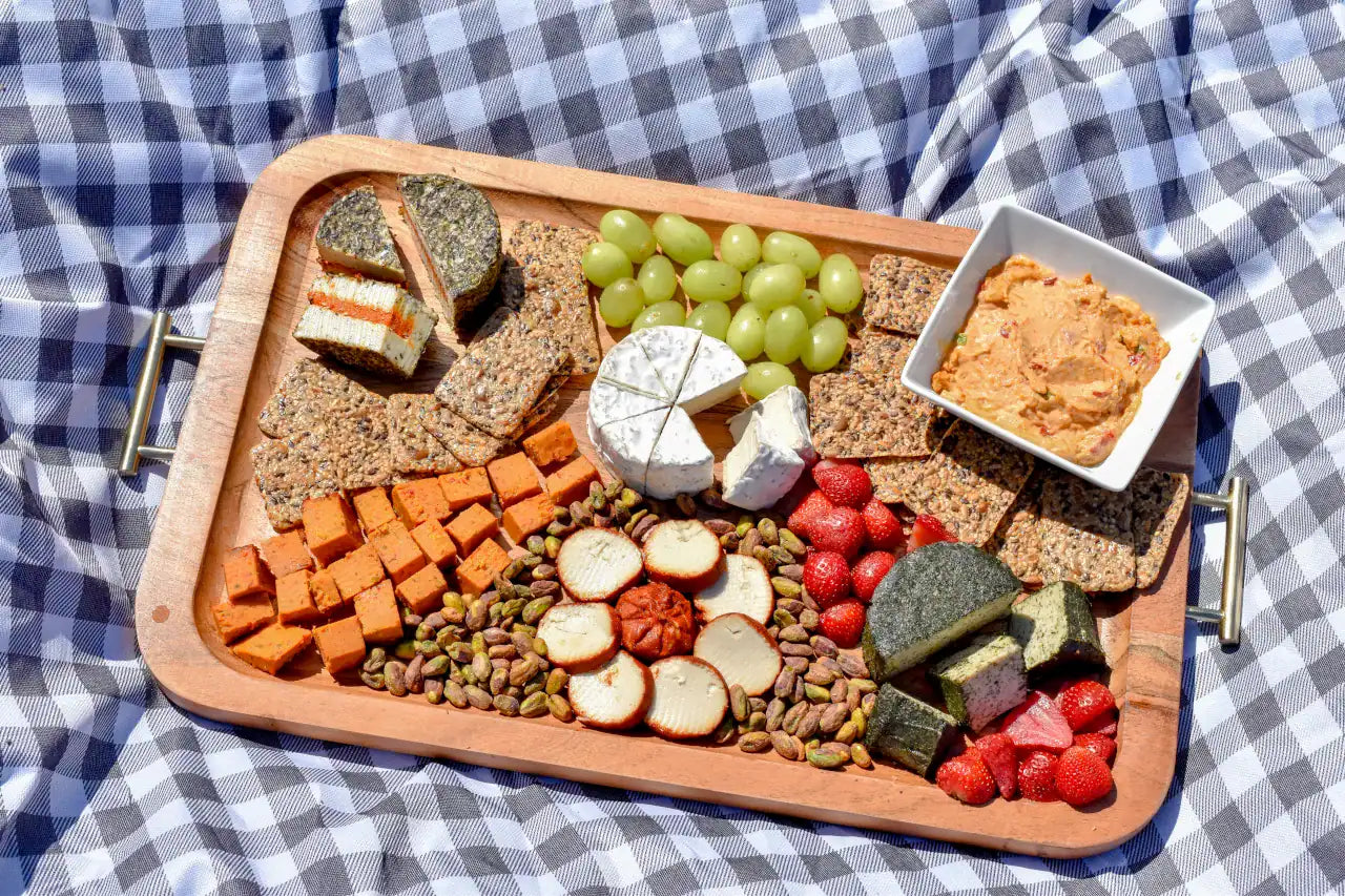 Wooden tray with assorted dairy-free cheeses and snacks on a checkered fabric background.