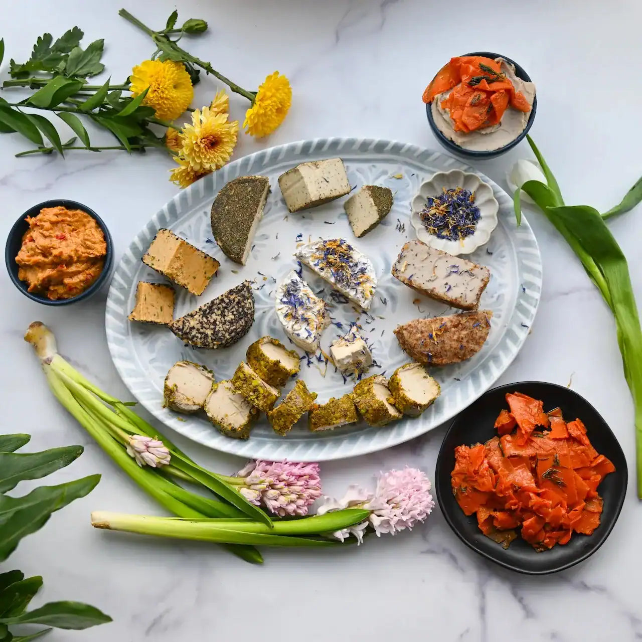 Assorted artisanal dairy-free cheeses on a marble surface with flowers and greenery from Rebel Cheese.