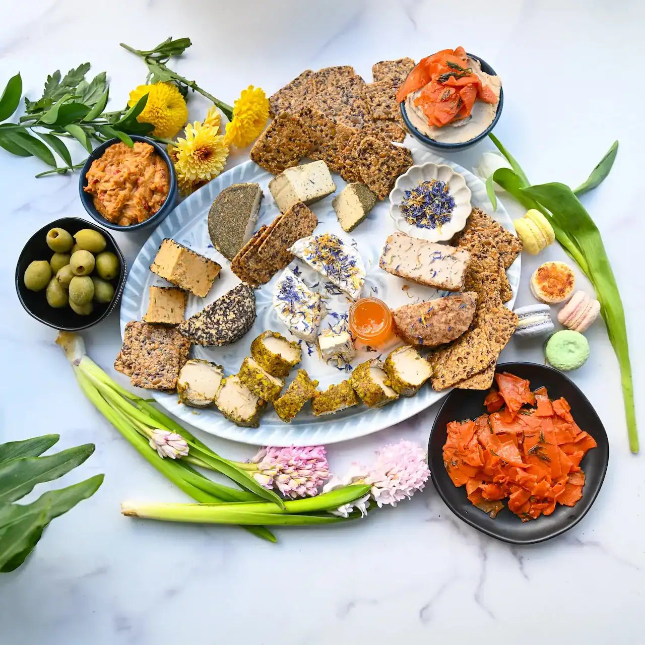 Assorted dairy-free cheeses and gluten-free crackers on a platter with flowers and greenery on a marble surface from Rebel Cheese.
