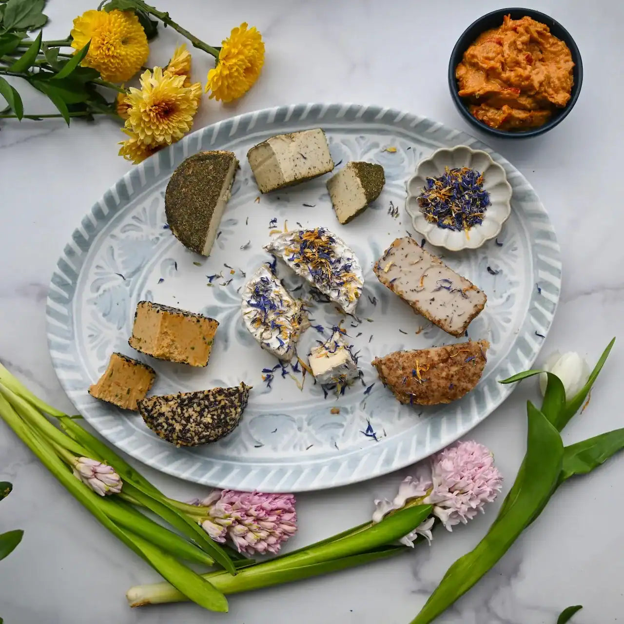 Platter of assorted dairy-free cheeses with flowers and a bowl of Pimento cheese on a marble surface from Rebel Cheese.