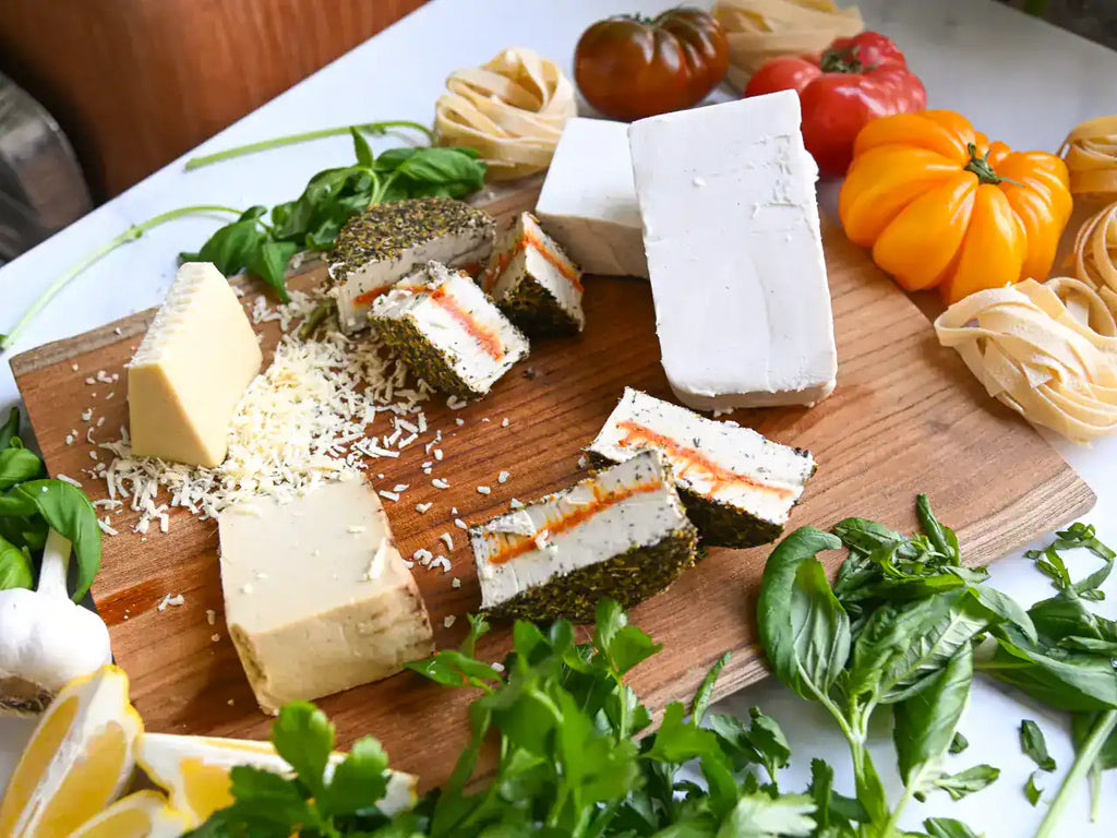 Six assorted dairy-free cheeses on a wooden cutting board with fresh basil and heirloom tomatoes along the edge of the board