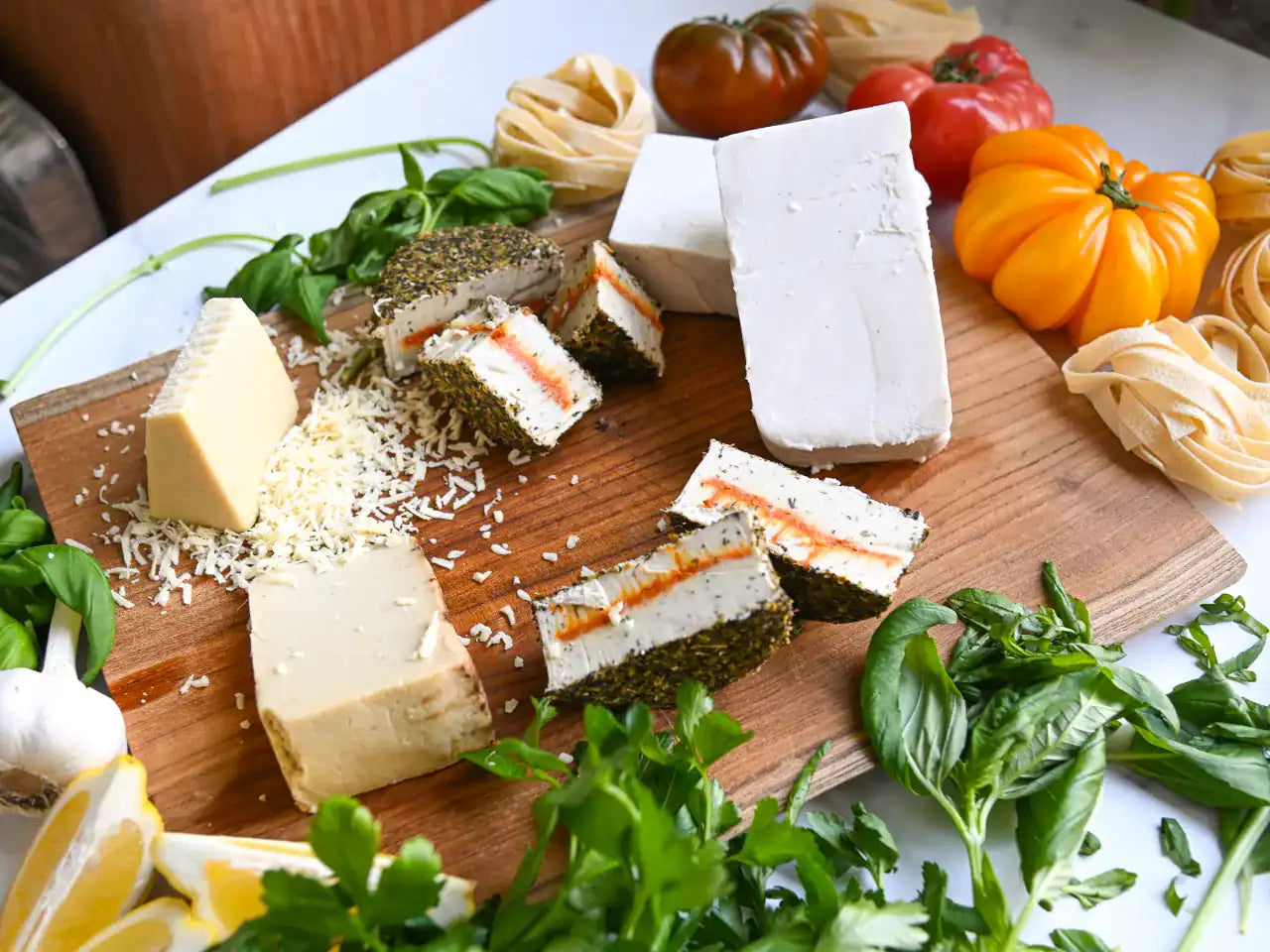 Six assorted dairy-free cheeses on a wooden cutting board with fresh basil and heirloom tomatoes along the edge of the board
