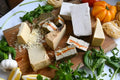Eight assorted dairy-free cheeses on a wooden cutting board with fresh basil and heirloom tomatoes along the edge of the board