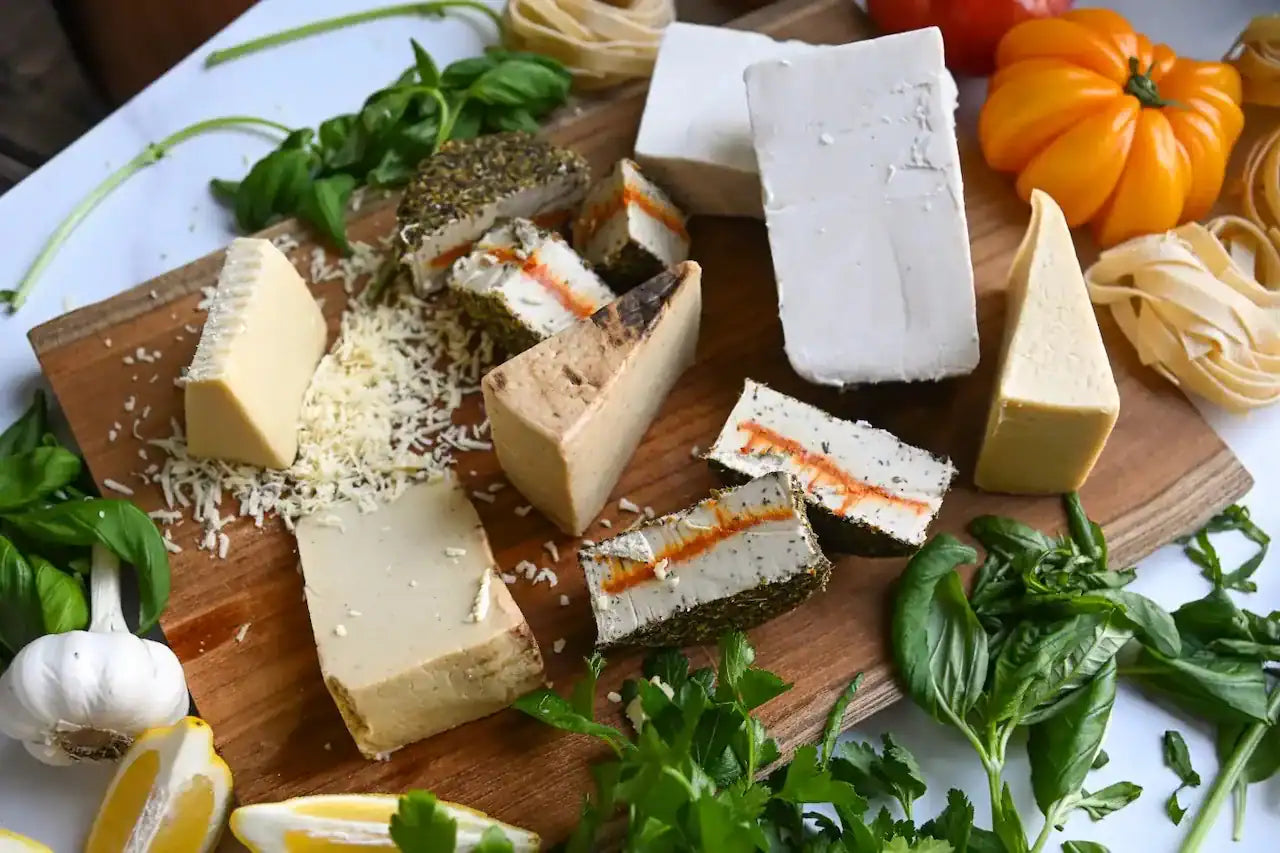 Eight assorted dairy-free cheeses on a wooden cutting board with fresh basil and heirloom tomatoes along the edge of the board