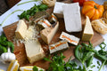 Eight assorted dairy-free cheeses on a wooden cutting board with fresh basil and heirloom tomatoes along the edge of the board