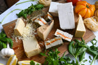 Eight assorted dairy-free cheeses on a wooden cutting board with fresh basil and heirloom tomatoes along the edge of the board